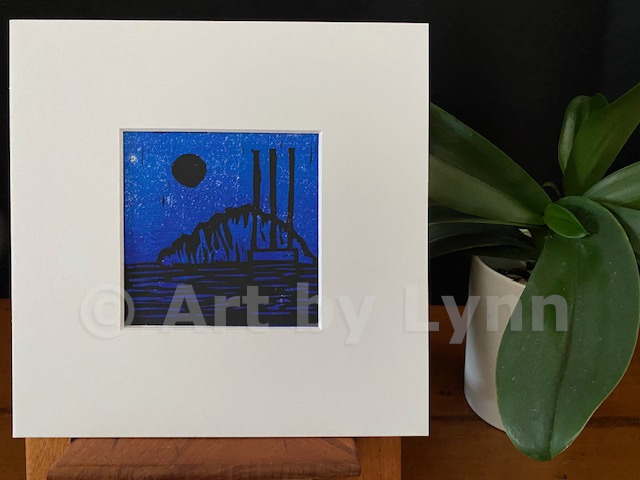 Framed relief print of the Rock in Morro Bay at night, next to a house plant.